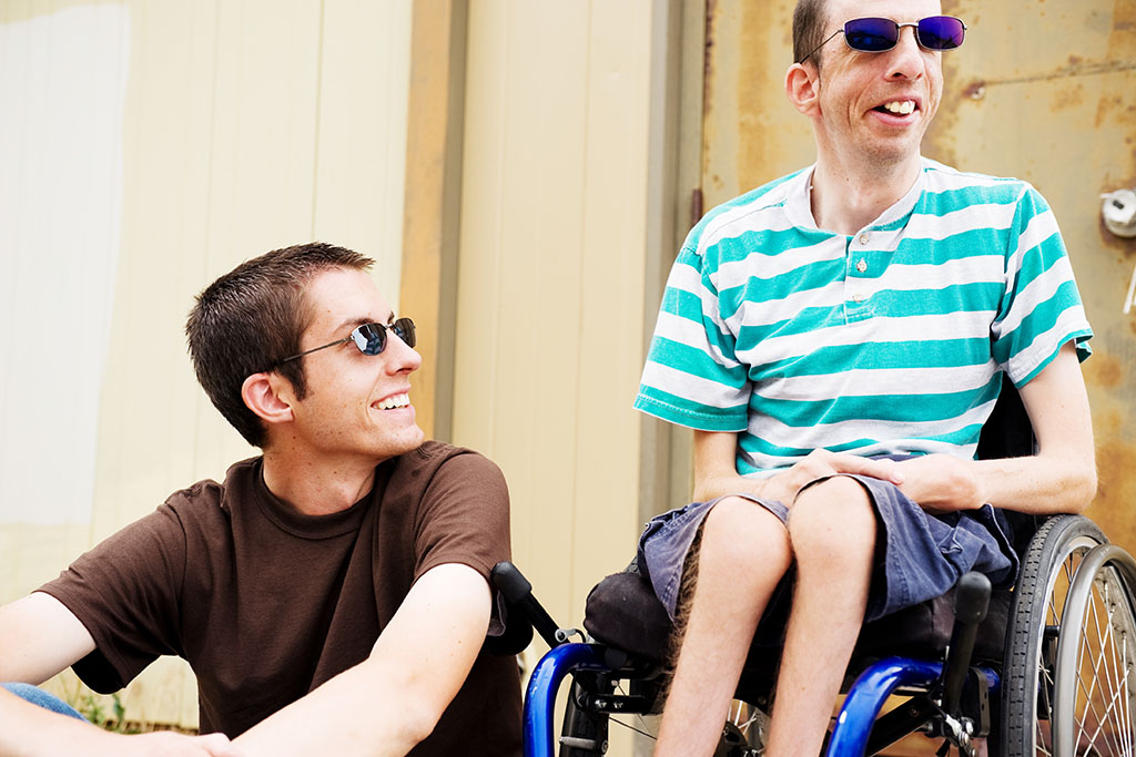 Two young men wearing sunglasses and summer clothing sit next to one another. The man on the right is in a wheelchair. Both are smiling.